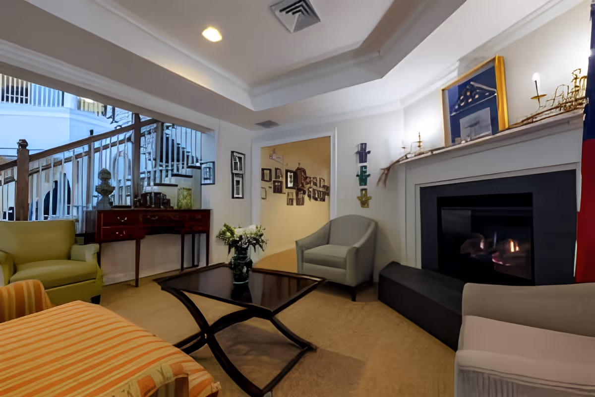 A cozy living room area with a fireplace, featuring a green armchair, a gray armchair, and a striped orange ottoman. A dark wooden coffee table with a vase of flowers sits in the center. The room has white walls with decorative wall art and a large mirror reflecting a staircase. The fireplace mantel is decorated with a framed picture and candle holders.