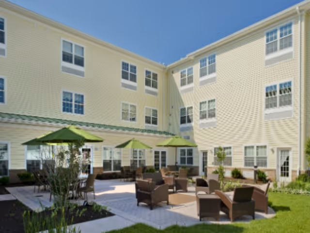 Outdoor courtyard area of a senior living facility with multiple brown wicker chairs and tables arranged on a paved patio. Several green umbrellas provide shade. The building surrounding the courtyard is three stories tall with numerous windows and light yellow siding. There is green grass and some landscaping around the patio.