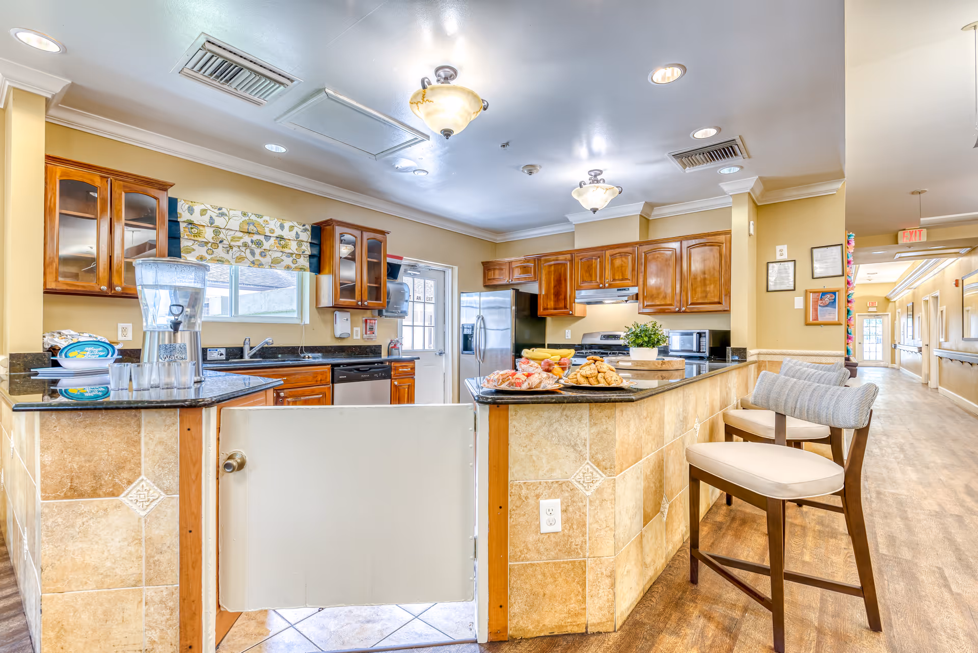 Bright communal kitchen with a central island topped with snacks, wooden cabinets, bar stools, and a hallway visible in the background.