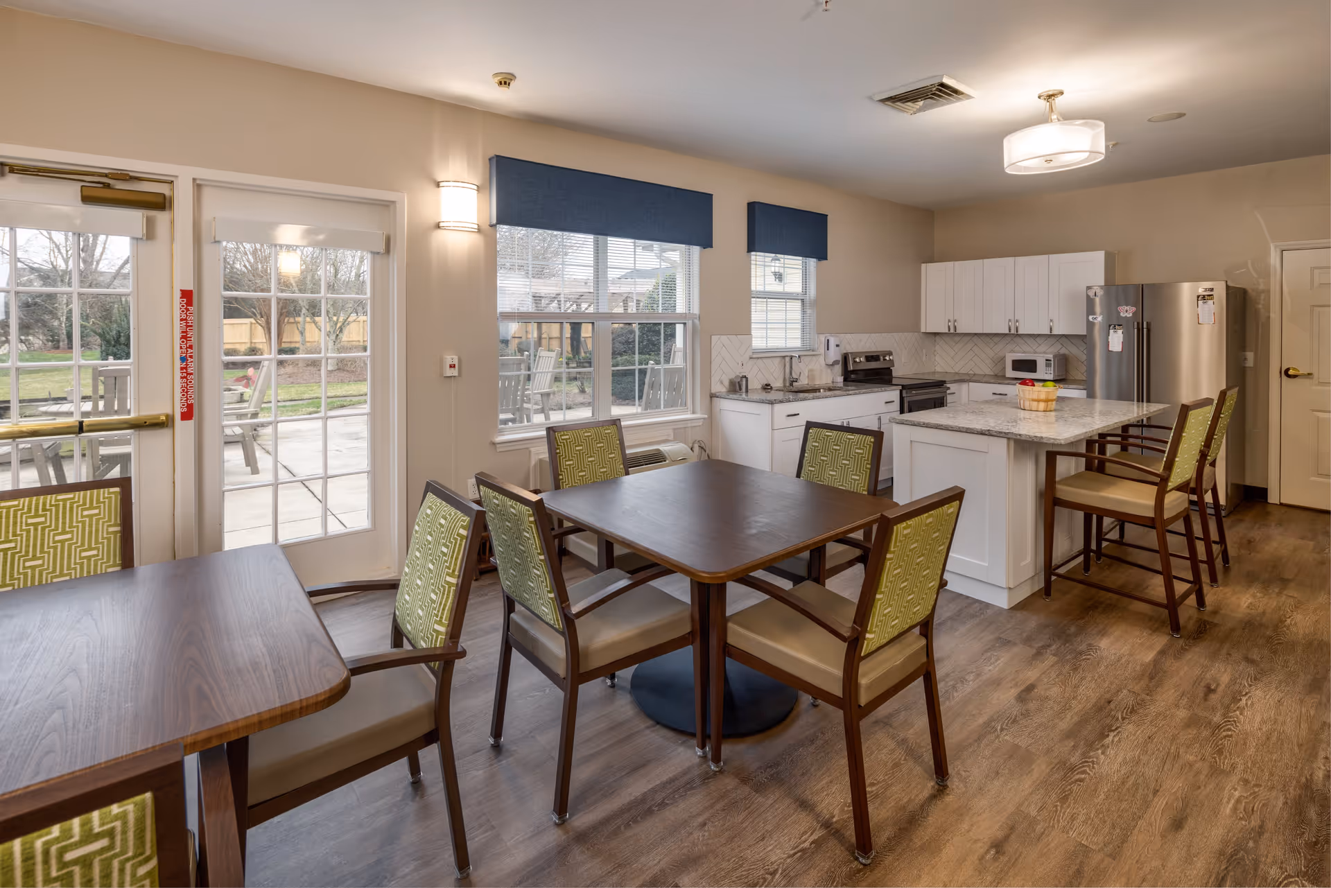 A bright and clean kitchen and dining area with wooden floors, white cabinets, a stainless steel refrigerator, a microwave, and a kitchen island with two chairs. There are two tables with green cushioned chairs, large windows with blue valances, and a glass door leading to an outdoor patio with chairs and tables.