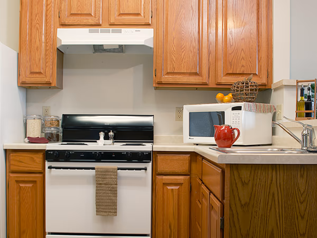 Compact kitchen with oak cabinets, a white stove and microwave, sink, and a red teapot on the counter.