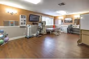 Spacious rehabilitation therapy room with treatment tables, exercise equipment, a wall-mounted TV, and hardwood floors.