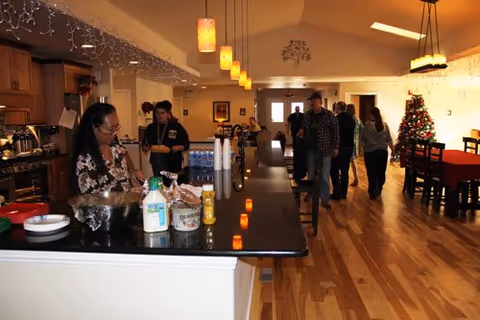 Interior view of a spacious assisted living facility with a kitchen island in the foreground where a woman is preparing food. Several people are walking and standing in the background near a decorated Christmas tree and dining tables. The room has wooden floors, pendant lights, and a high ceiling.