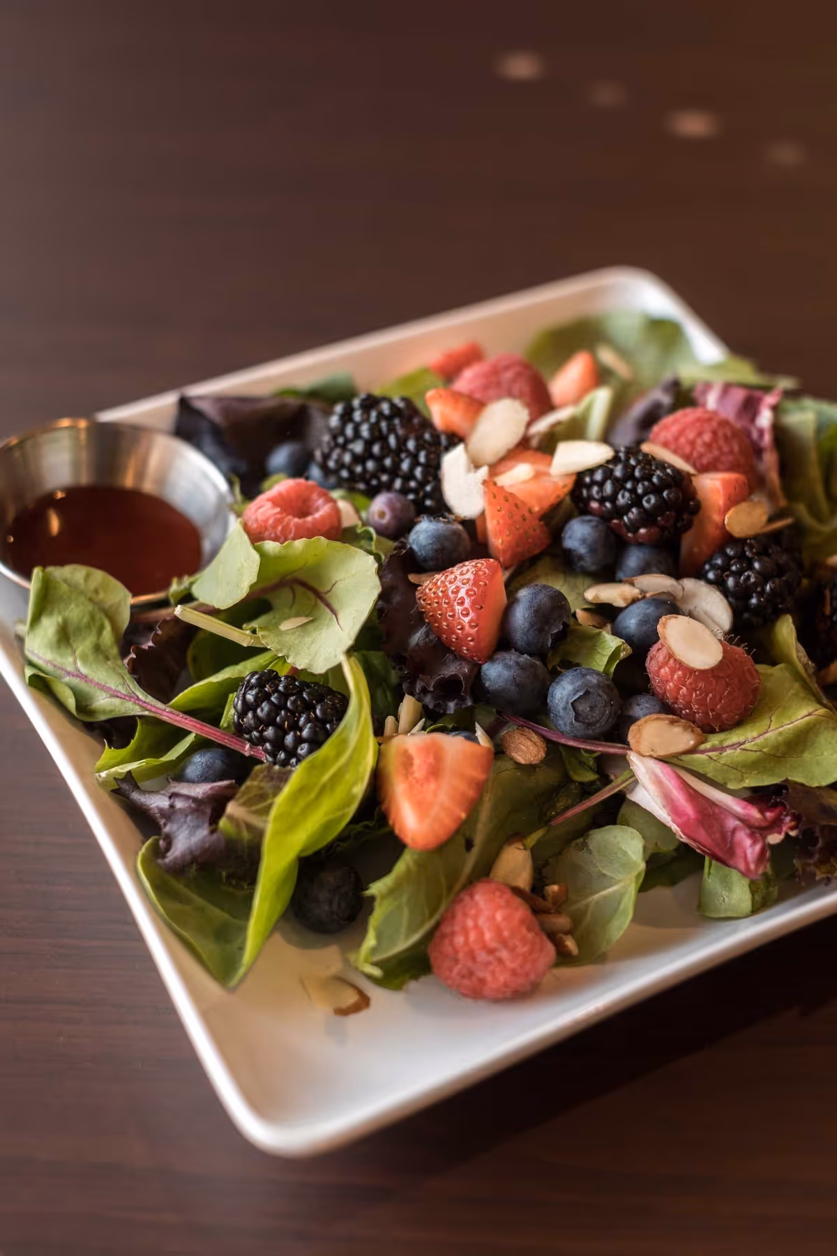 A fresh salad with mixed greens, blackberries, raspberries, blueberries, sliced strawberries, and almond slices on a white square plate with a small metal cup of dressing on the side.