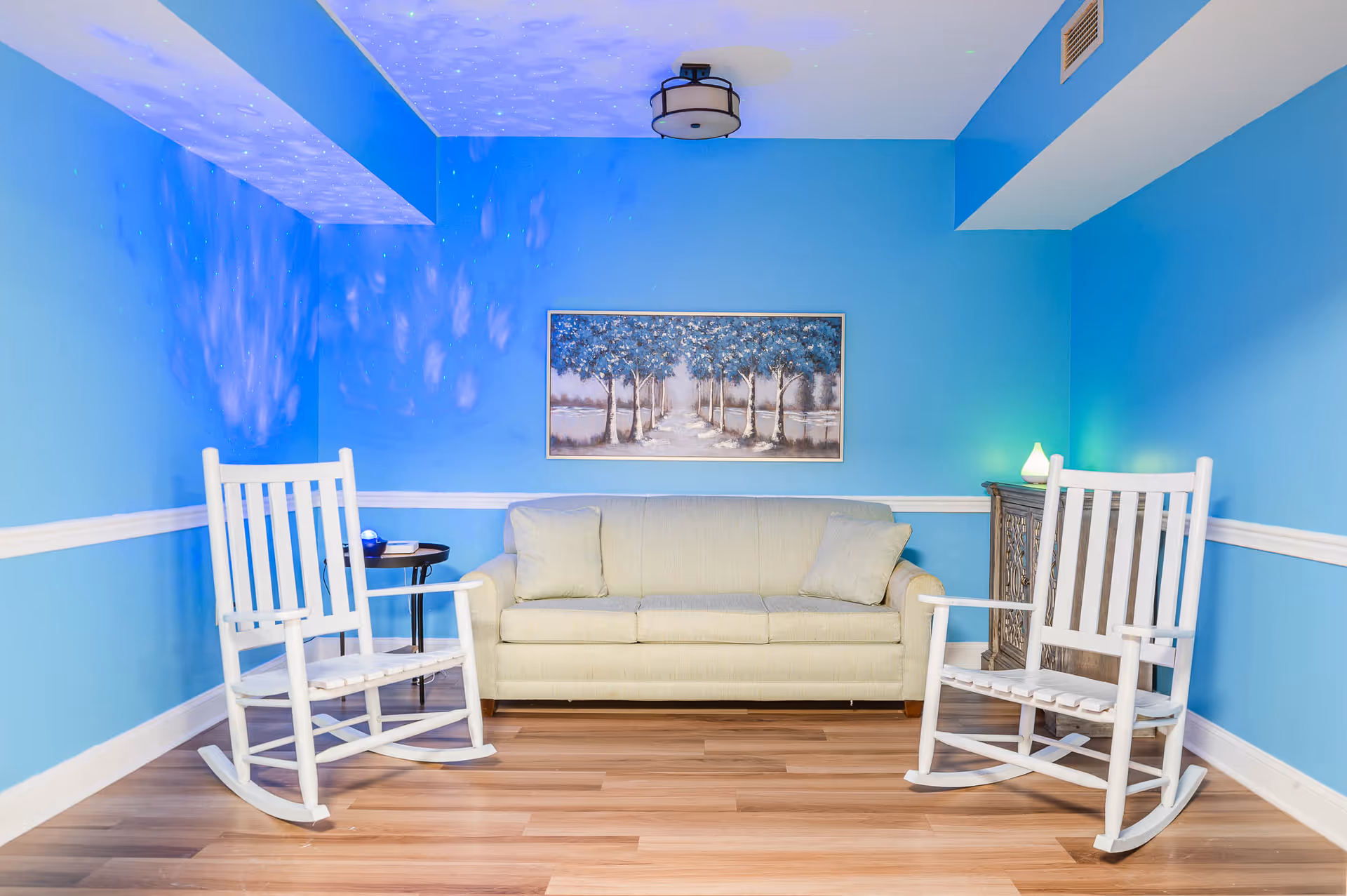 A cozy living room with light blue walls and wooden flooring. The room features a beige sofa with two cushions, flanked by two white wooden rocking chairs. A small round side table with a blue decorative item is placed next to the left rocking chair. On the right side, there is a wooden cabinet with a small lamp emitting a soft green light. Above the sofa, a framed painting of a tree-lined path hangs on the wall. The ceiling light fixture is modern and round, and there are subtle light patterns projected on the walls and ceiling.