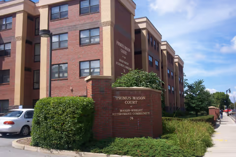 Exterior view of a multi-story brick building with beige accents, identified as Primus Mason Court at Mason-Wright Retirement Community. The building is surrounded by greenery including bushes and trees, with a sidewalk and parked cars visible nearby under a partly cloudy sky.
