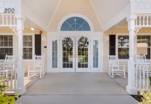 Covered front porch with double glass doors adorned with floral wreaths, flanked by white rocking chairs and decorative columns.