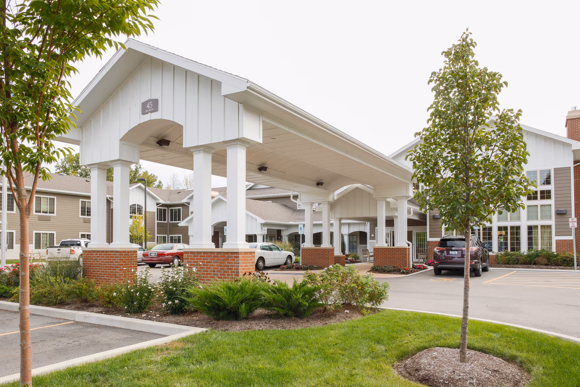 Exterior view of The Village at Mill Landing senior living facility showing a covered entrance with white pillars and brick bases, surrounded by landscaped greenery and a parking area with several cars.