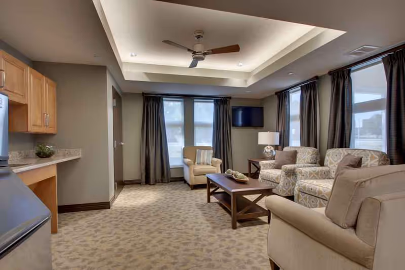 Cozy common living room with upholstered armchairs, a coffee table, a wall-mounted TV, and windows beneath a tray ceiling with a ceiling fan.