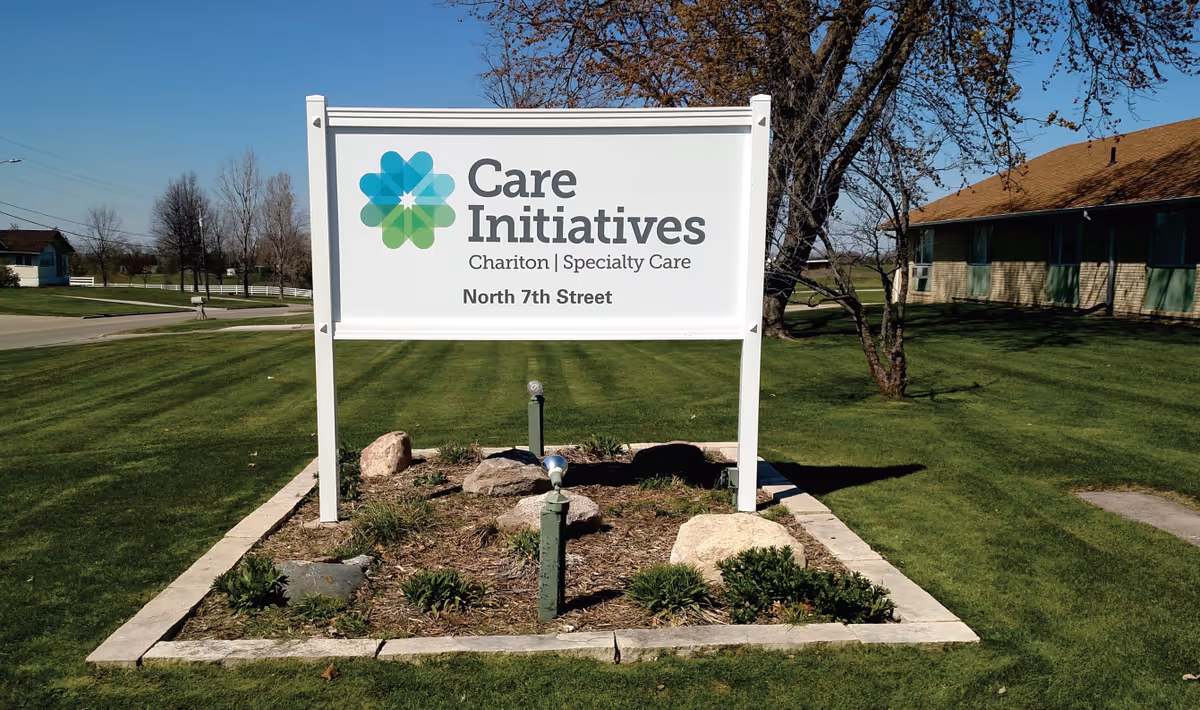 Outdoor view of a white sign for Care Initiatives Chariton Specialty Care located on North 7th Street, surrounded by a small landscaped area with rocks and plants, with a building and trees in the background under a clear blue sky.