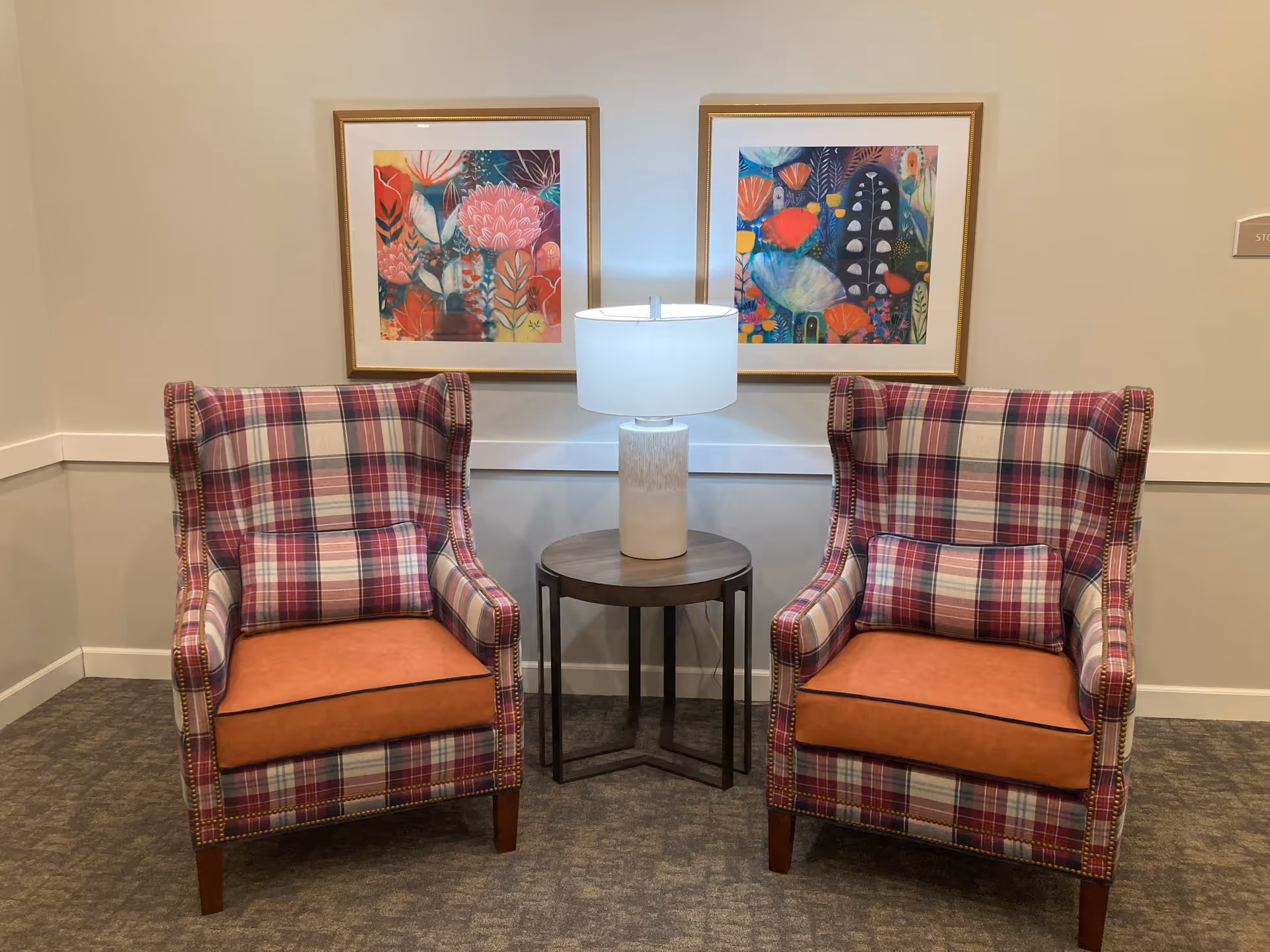 Two plaid armchairs with orange seat cushions flank a small round table and lamp beneath two colorful framed prints on a beige wall.