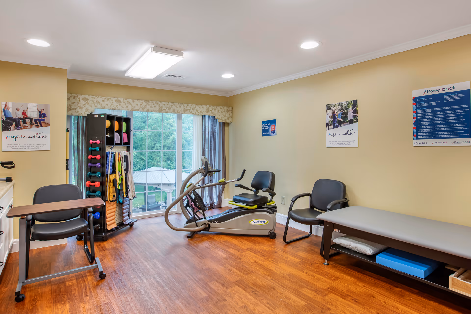A small exercise room with wooden flooring, a NuStep recumbent cross trainer, a rack holding various exercise equipment like dumbbells and resistance bands, two chairs, a table, and a padded bench. The room has large windows with a view of greenery outside and posters on the walls promoting physical activity.