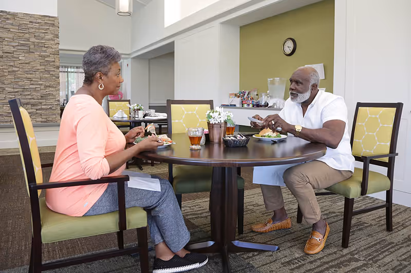Two elderly individuals sitting at a round dining table in a well-lit room, eating a meal and engaging in conversation. The room has a modern design with a stone accent wall and green cushioned chairs with yellow patterned backs.