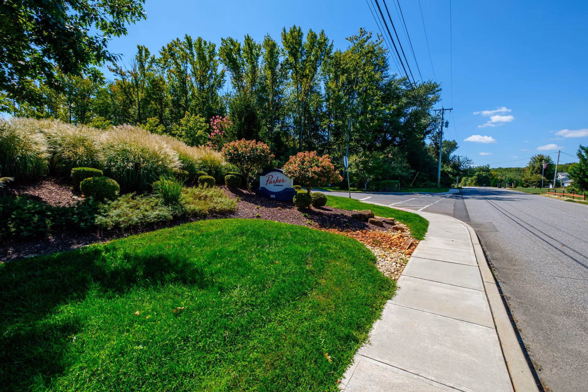 Landscaped entrance with a Parker sign beside a sidewalk and road, framed by trees and a blue sky.
