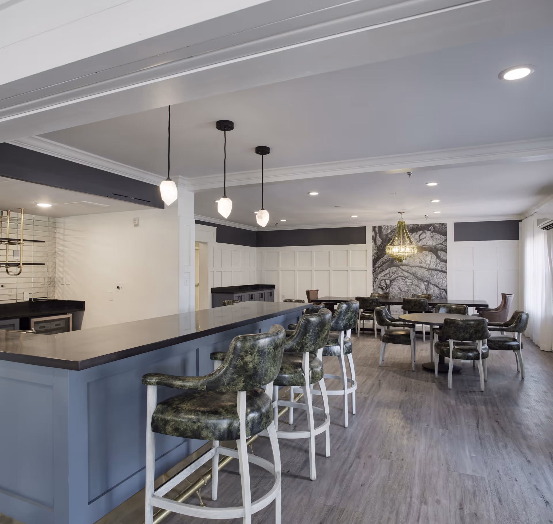 A modern communal dining and lounge area with a long bar counter and several green cushioned bar stools. The room features round tables with matching chairs, a chandelier, and a large black and white tree mural on the far wall. The space has wood flooring, white paneled walls, and pendant lights hanging above the bar.