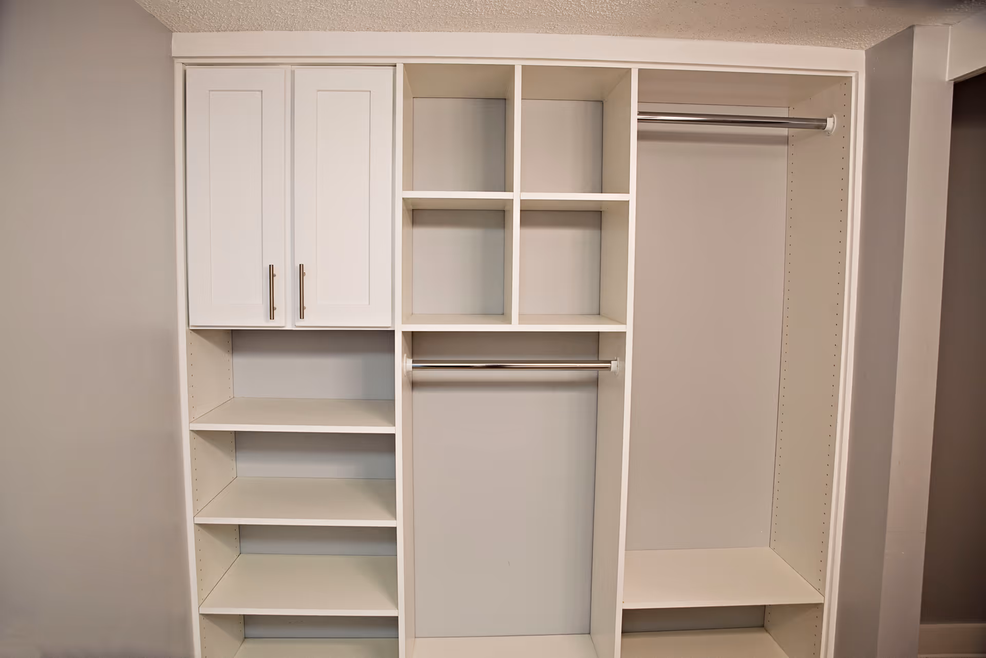 Empty white built-in closet organizer with shelves, cabinets, cubbies, and hanging rods against a beige wall.