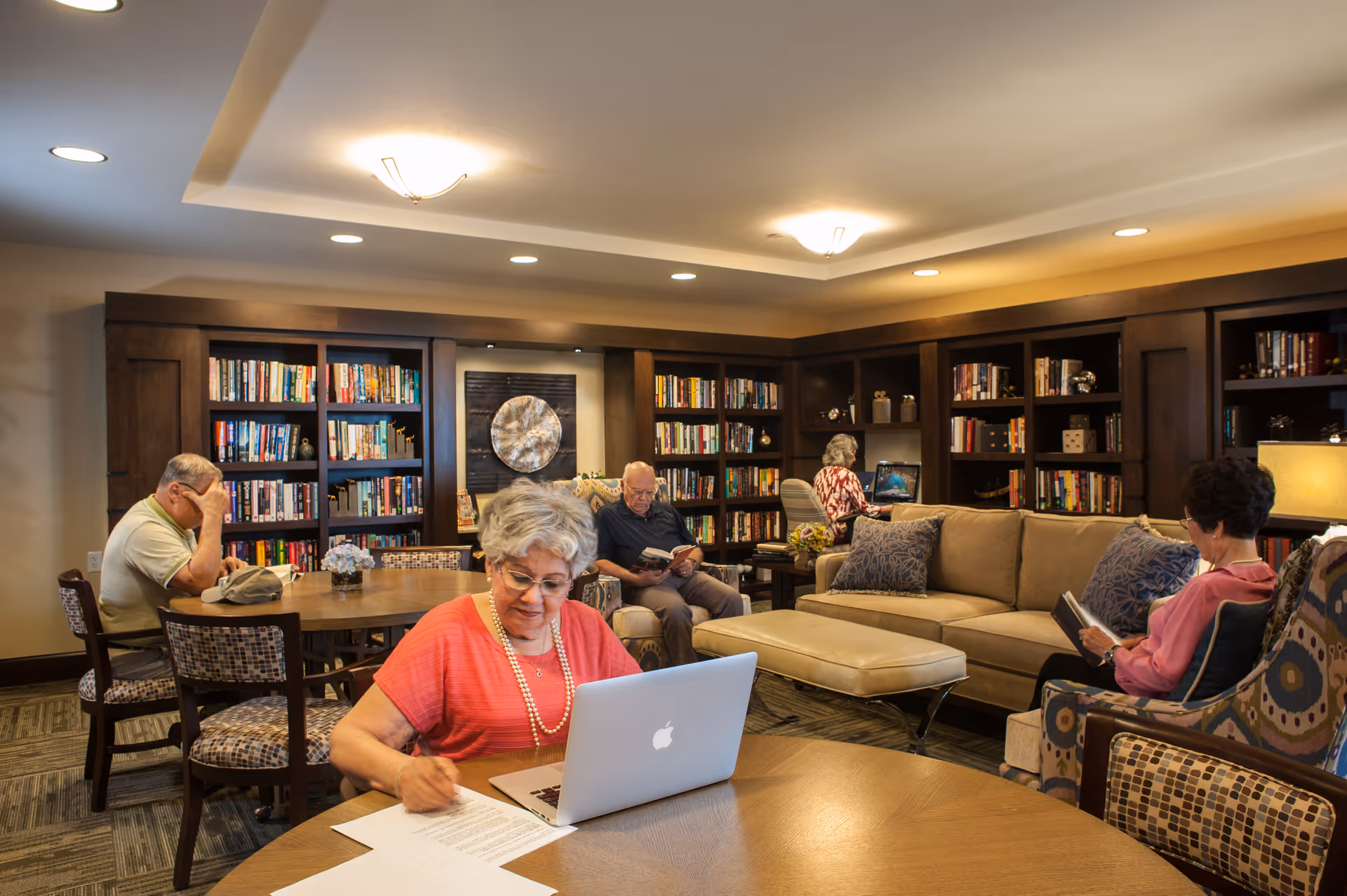 A cozy library room in a senior living facility with several elderly people engaged in different activities. One woman in the foreground is working on a laptop and writing on papers at a round table. In the background, a man is reading a book while sitting on a chair, another woman is using a laptop at a desk, and another woman is reading a book while seated in an armchair. The room has wooden bookshelves filled with books and warm lighting.