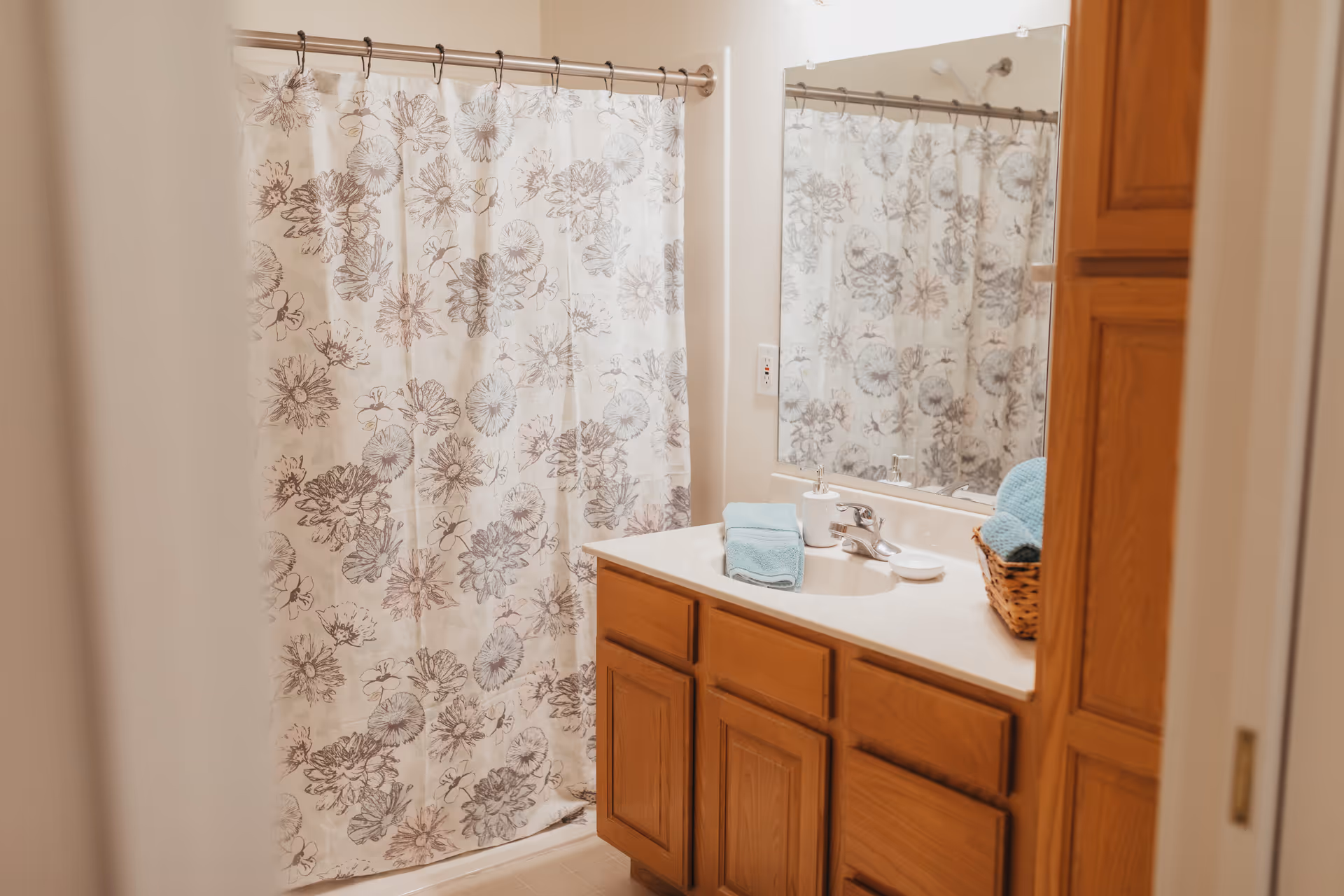 Bathroom with a floral patterned shower curtain, wooden cabinets under a white countertop with a sink, a mirror above the sink, and a basket with folded blue towels on the counter.
