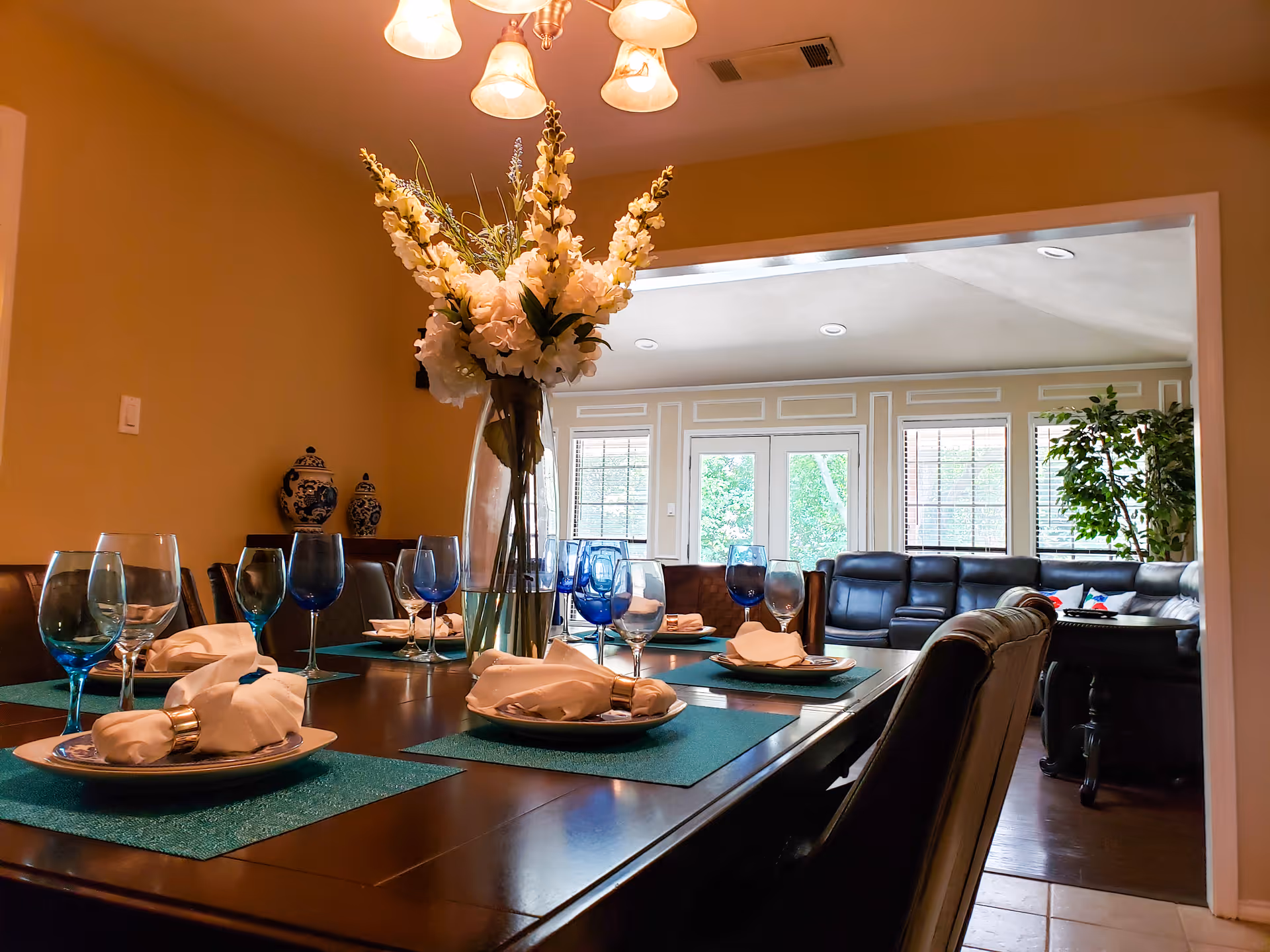 A formal dining table set with blue wine glasses, white napkins, and a tall vase of white flowers opening into a bright living room.