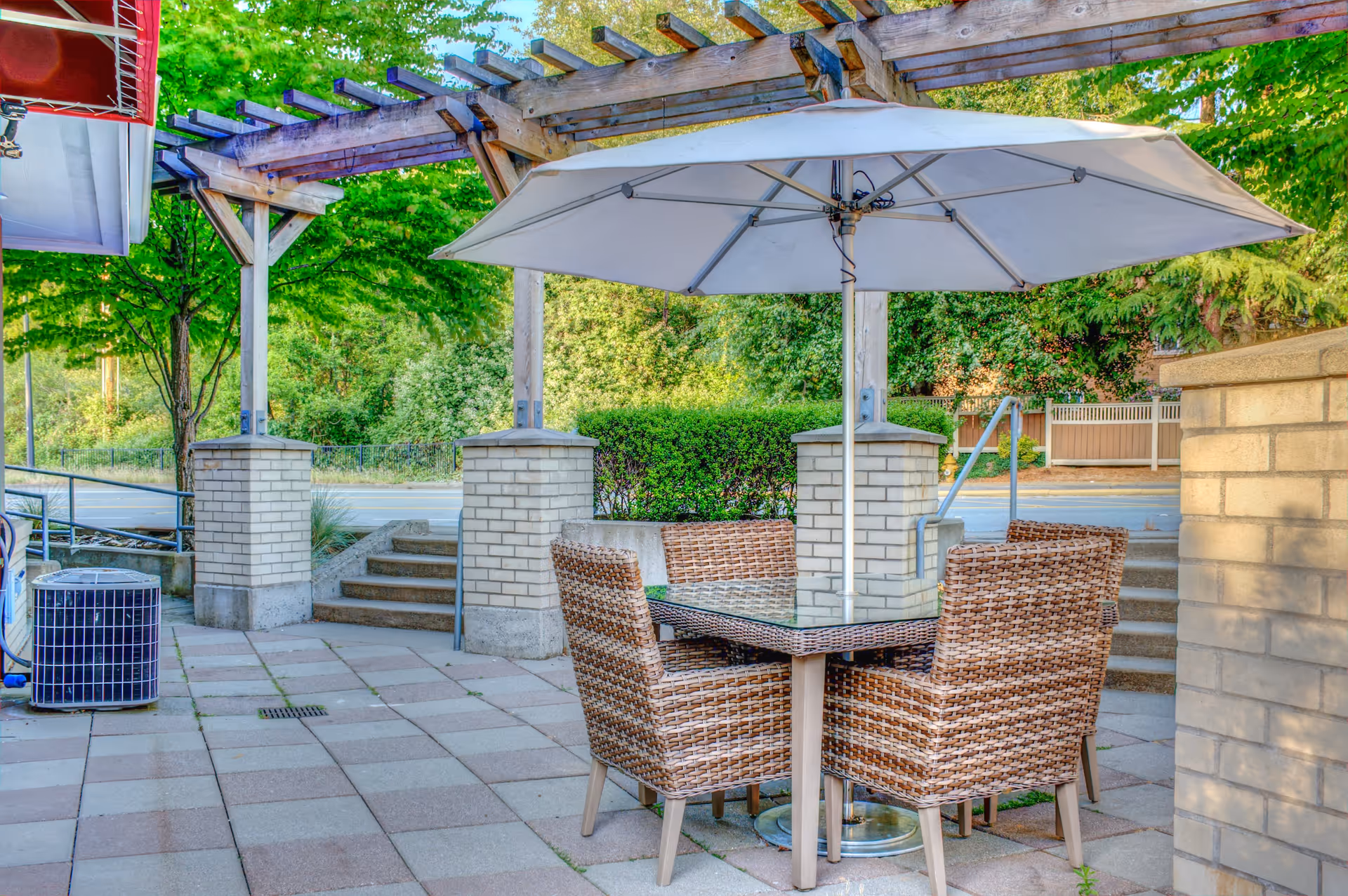 Outdoor patio area with a square glass-top table surrounded by four wicker chairs under a large beige umbrella. The patio is paved with square tiles and features brick pillars supporting a wooden pergola. Green trees and bushes are visible in the background along with steps leading up to a street.