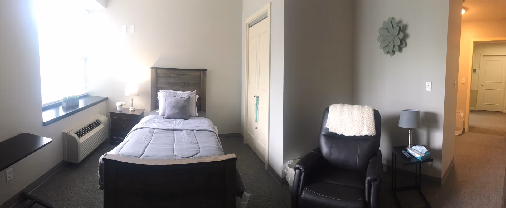 A small, neatly arranged bedroom with a single bed made with gray bedding and a pillow. Next to the bed is a nightstand with a lamp and a small plant on the windowsill. A black recliner chair with a white throw blanket is positioned near a small side table with a lamp and tissues. The walls are painted light gray, and there is a decorative flower-shaped wall hanging. A closet with closed doors is visible, and the room opens into a hallway.