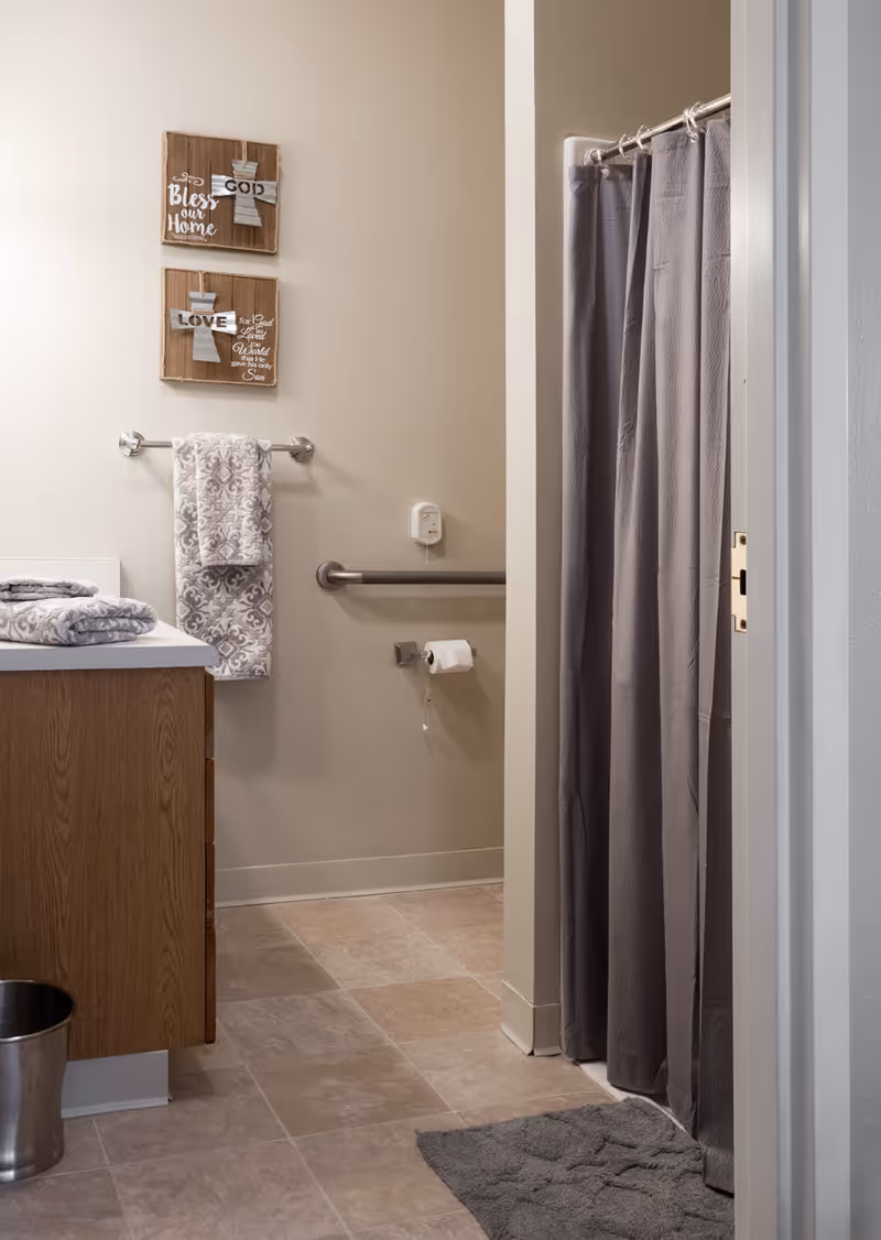 A bathroom with beige walls and tiled floor featuring a wooden vanity with folded towels on the counter, a metal trash can, a towel rack with a patterned towel, a toilet paper holder with a roll, a grab bar on the wall, and a shower with a gray curtain. Two decorative wooden plaques with crosses and inspirational text hang on the wall.