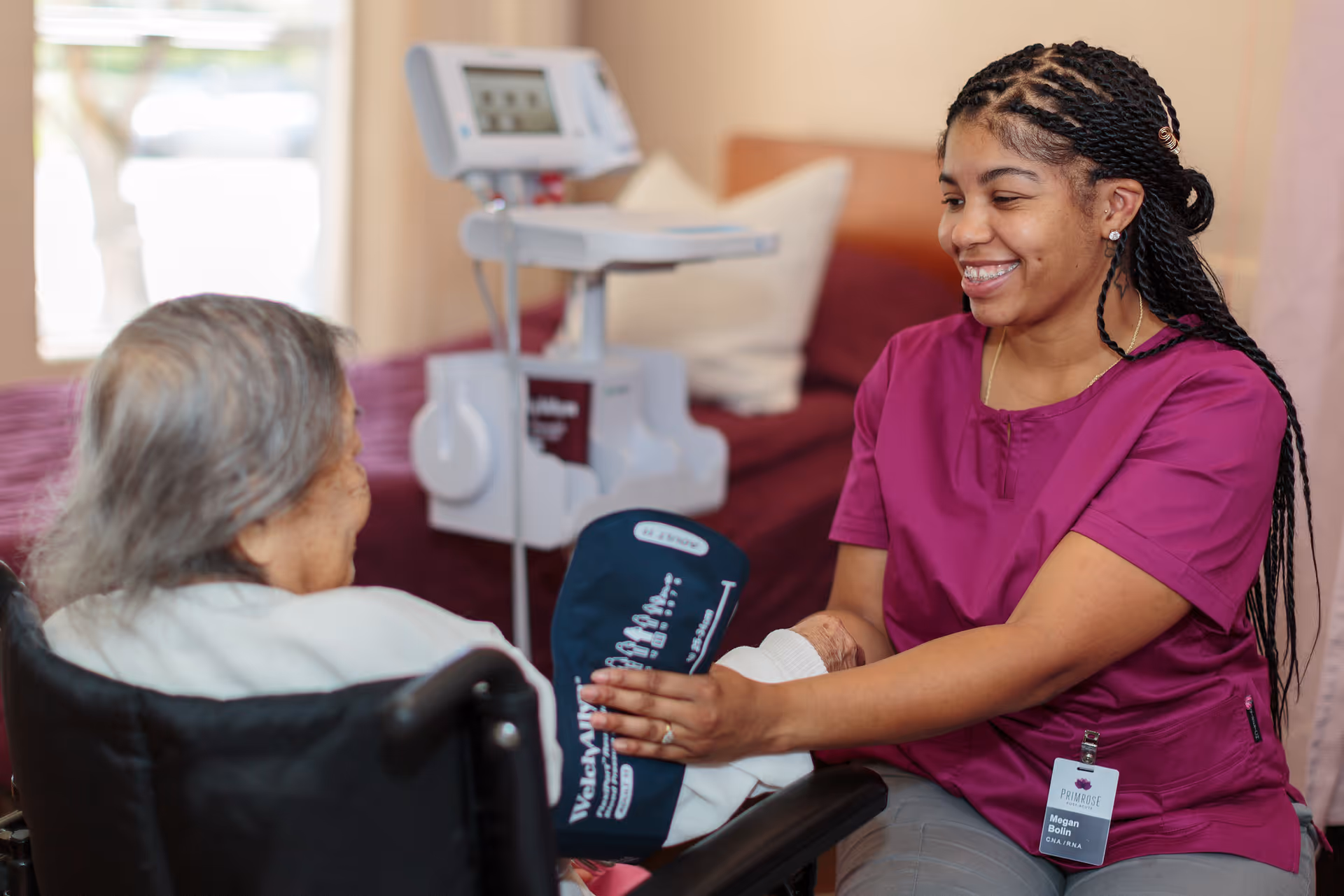 A healthcare worker in a purple uniform is smiling while taking the blood pressure of an elderly person seated in a wheelchair inside a room with a bed and medical equipment in the background.