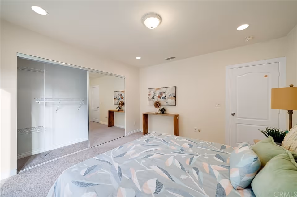 A bedroom with a bed covered in a patterned comforter and several pillows. There is a wooden console table against the wall with decorative items and a framed abstract painting above it. The room has a carpeted floor, a closet with sliding mirrored doors, and a closed white door. A lamp and a small plant are visible on the right side of the bed.