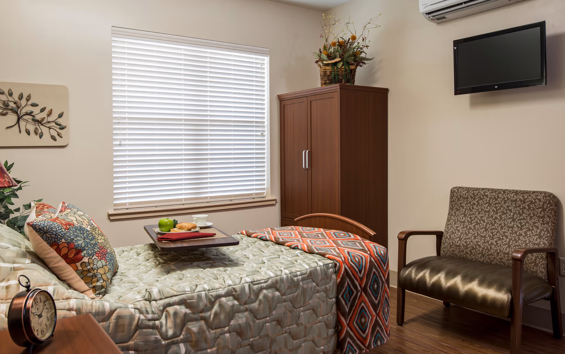 A furnished senior care bedroom with a made bed holding a meal tray, decorative pillows and throw, a wooden armoire, armchair, and wall-mounted TV.