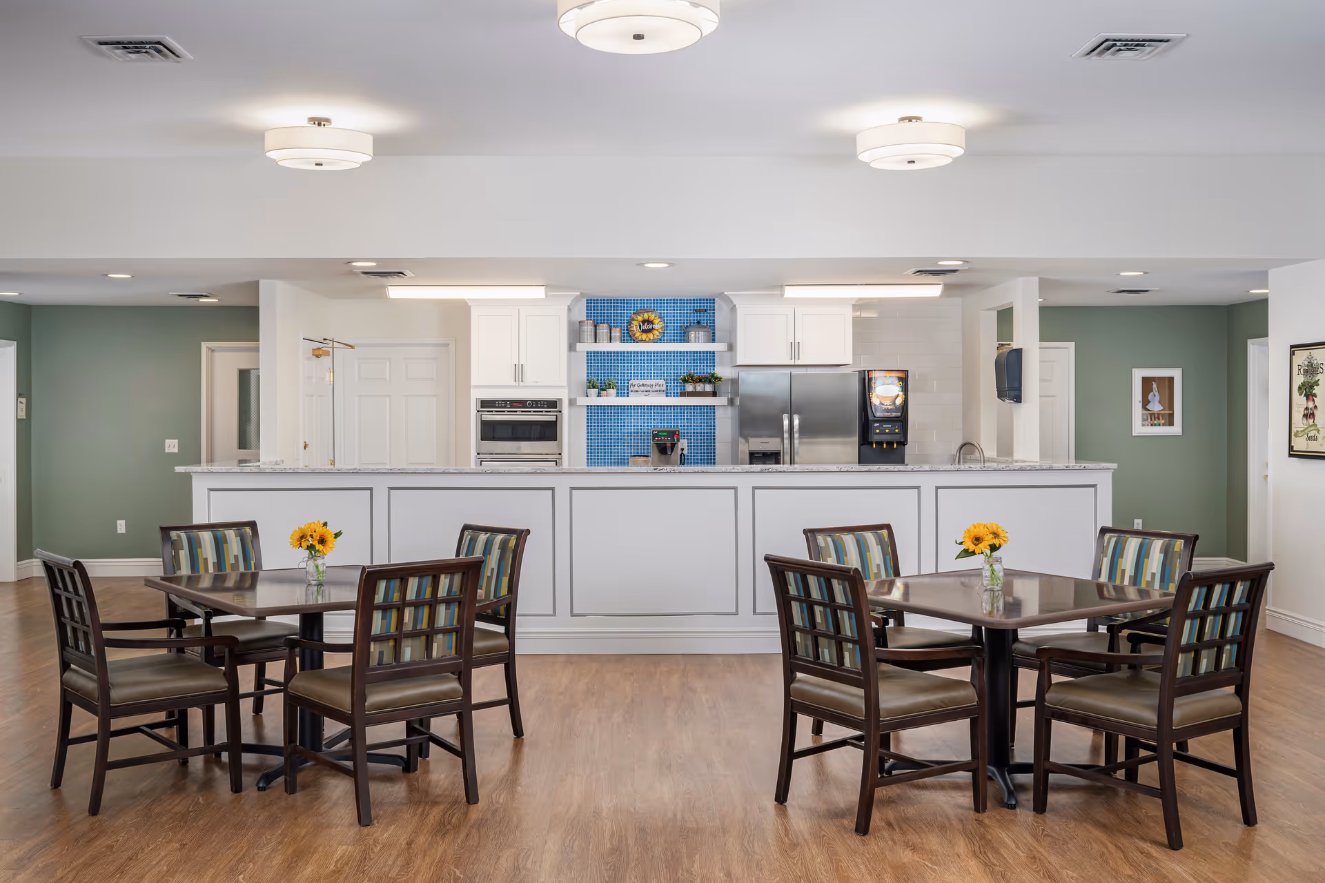 A bright and clean dining area in a senior living facility with two square tables, each surrounded by four chairs with patterned cushions. Each table has a small vase with yellow flowers. In the background, there is a kitchen counter with white cabinetry, a stainless steel refrigerator, a built-in oven, and a beverage dispenser. The walls are painted green and white, and the floor is wood.