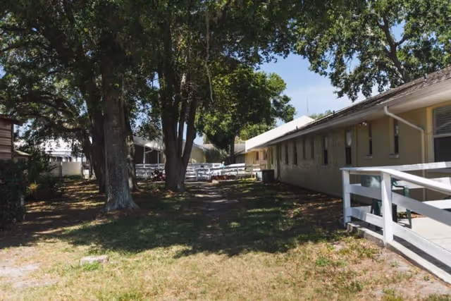 Side view of a single-story senior living building with a white fence and ramp, shaded yard and large trees.