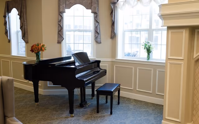 A black grand piano with a matching bench sits in a well-lit room with three large windows adorned with valances. There are flower arrangements on the piano and on the window sill. The room has light-colored walls with decorative paneling and a carpeted floor.
