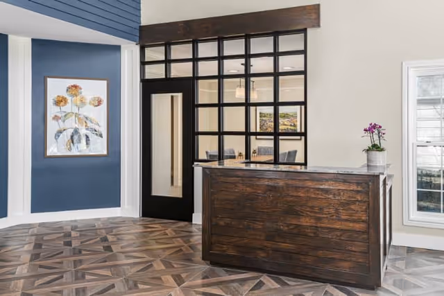 Reception area with a dark wooden front desk, a potted orchid on the desk, a large window with black grid framing behind the desk, a door with a glass panel, blue accent wall with a framed floral painting, and patterned wooden flooring.