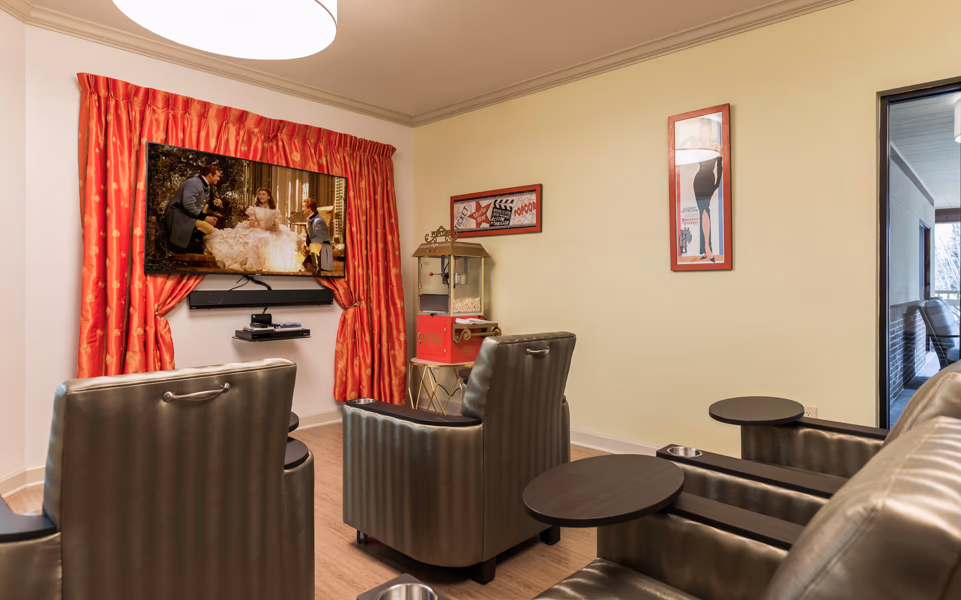 A cozy living room area with three dark leather recliner chairs facing a wall-mounted flat-screen TV playing a scene from a movie. The TV is framed by red curtains. There is a popcorn machine on a stand next to the TV, and two framed movie-themed posters hang on the light yellow wall. A glass door on the right leads to an outdoor patio area.