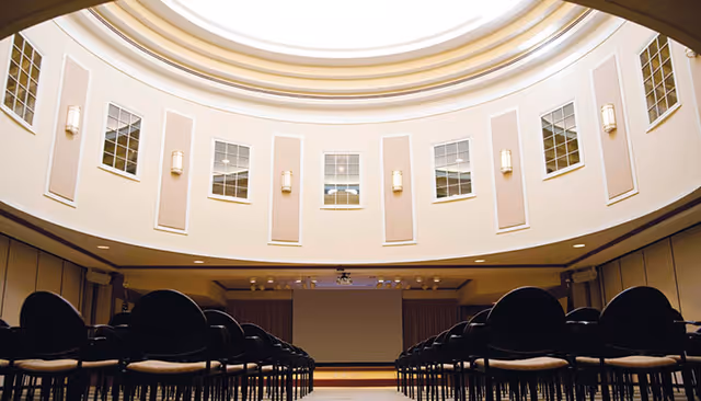 Interior view of a large auditorium or conference room with rows of black chairs facing a stage with a blank screen. The ceiling is high and circular with multiple windows and wall lights around the upper walls.
