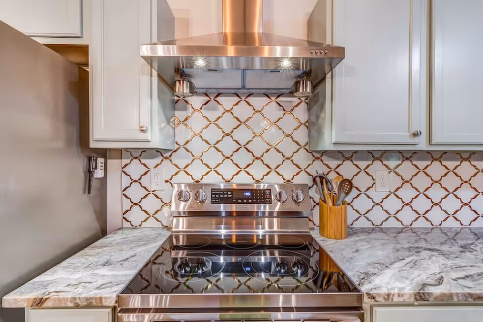 Modern kitchen stove with a stainless steel range hood, white cabinets, marble countertops, and a decorative tile backsplash with a geometric pattern. A wooden utensil holder with kitchen tools is placed on the right side of the stove.