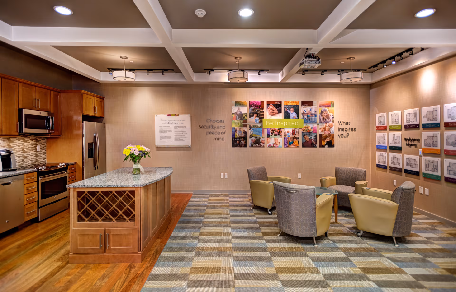 A modern interior room featuring a kitchen area with wooden cabinets, stainless steel appliances, and a granite countertop island with a vase of flowers. Adjacent to the kitchen is a seating area with four cushioned chairs arranged around a small glass table. The walls display colorful inspirational posters and floor plan layouts. The room has a patterned carpet and a coffered ceiling with recessed lighting and hanging light fixtures.