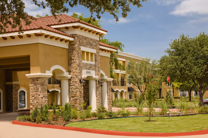 Exterior view of a senior living facility building with stone and stucco facade, red tile roof, columns at the entrance, landscaped greenery, and trees under a partly cloudy sky.