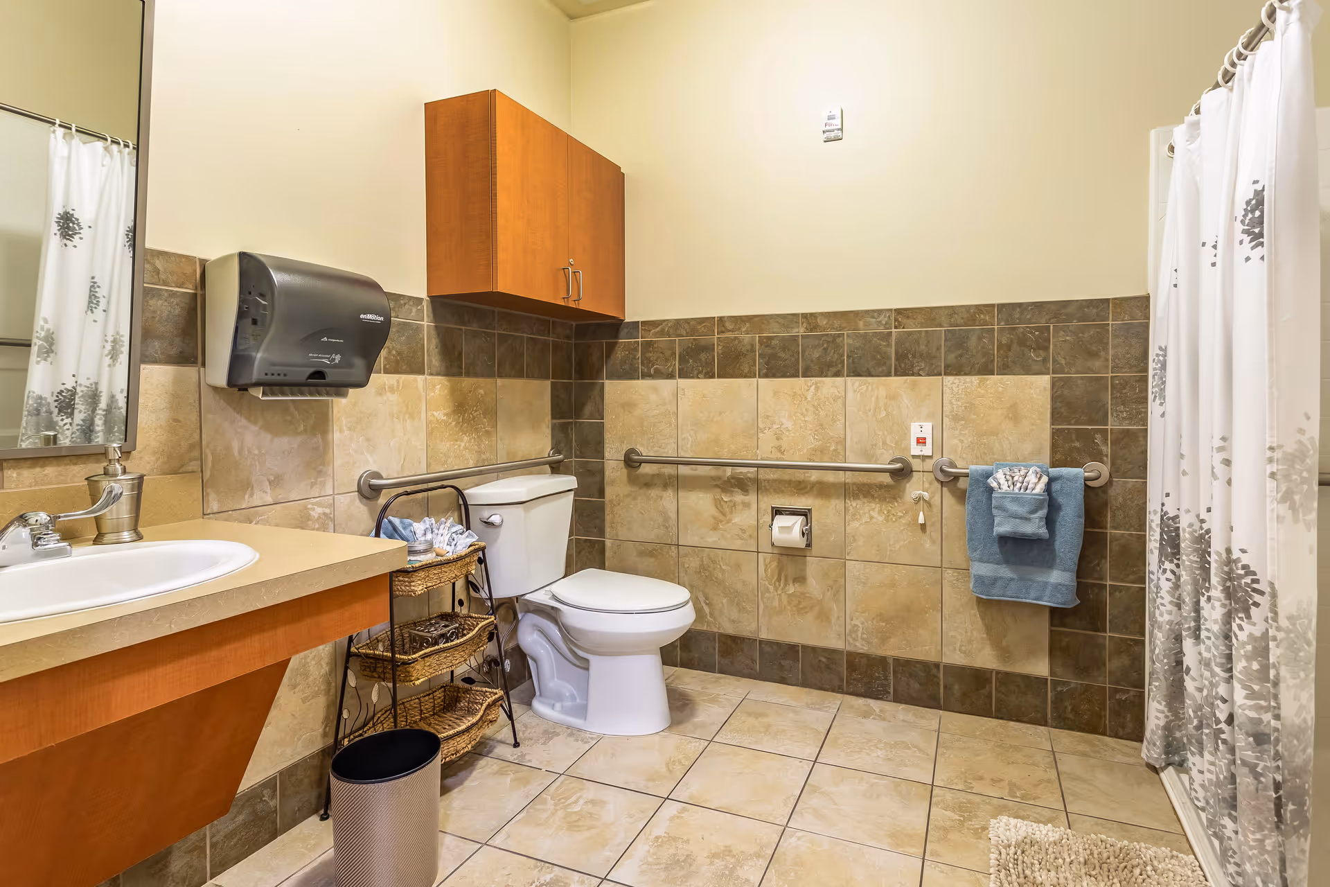 A clean and accessible bathroom with beige and brown tiled walls and floor. The room features a white toilet with grab bars on both sides, a sink with a countertop, a wall-mounted paper towel dispenser, a wooden cabinet above the toilet, a small metal rack with towels, and a shower area with a white curtain decorated with gray patterns.