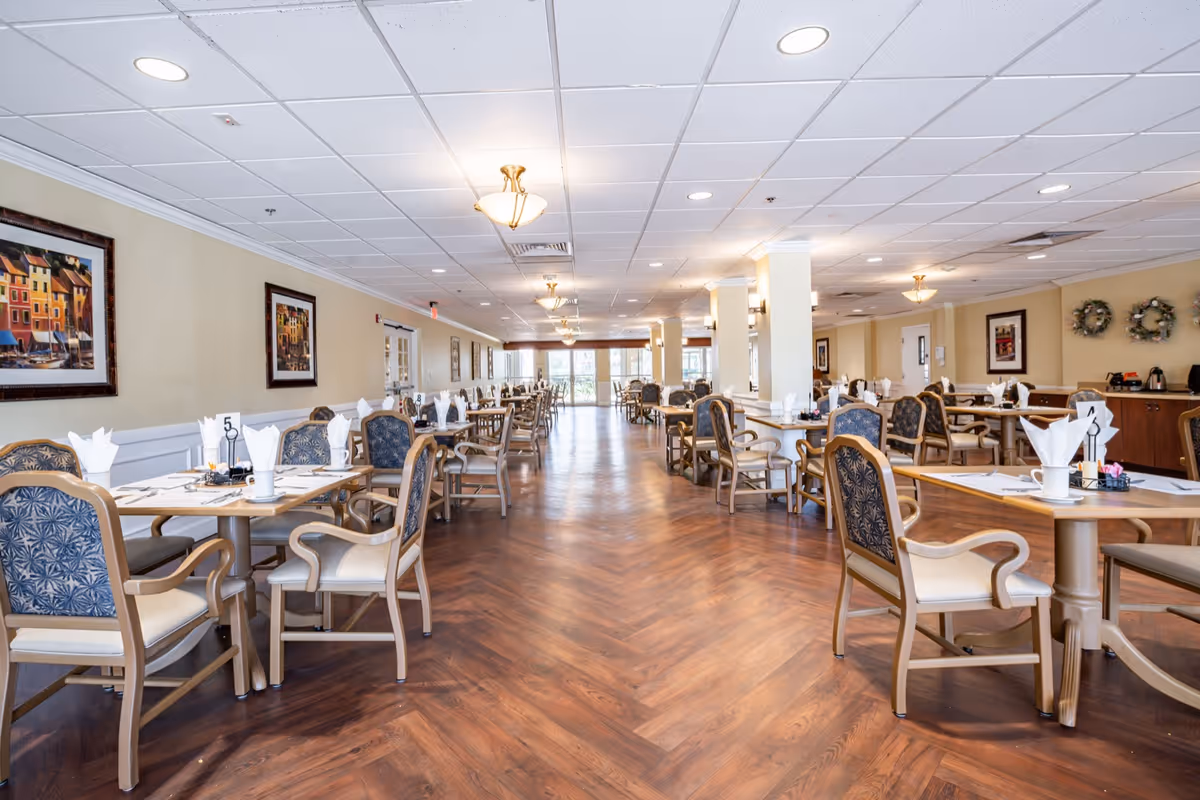 Spacious, well-lit communal dining room with multiple neatly set tables and chairs arranged along a wood-patterned floor.