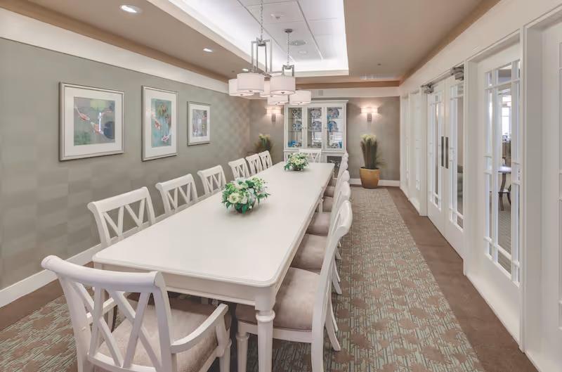 A long white dining table with twelve matching white chairs arranged around it in a well-lit dining room. The table has two floral centerpieces. The room features patterned carpet, light-colored walls with three framed artworks, a ceiling light fixture, a glass-front cabinet at the far end, and two potted plants on either side of the cabinet. There are white double doors with glass panels on the right side of the room.