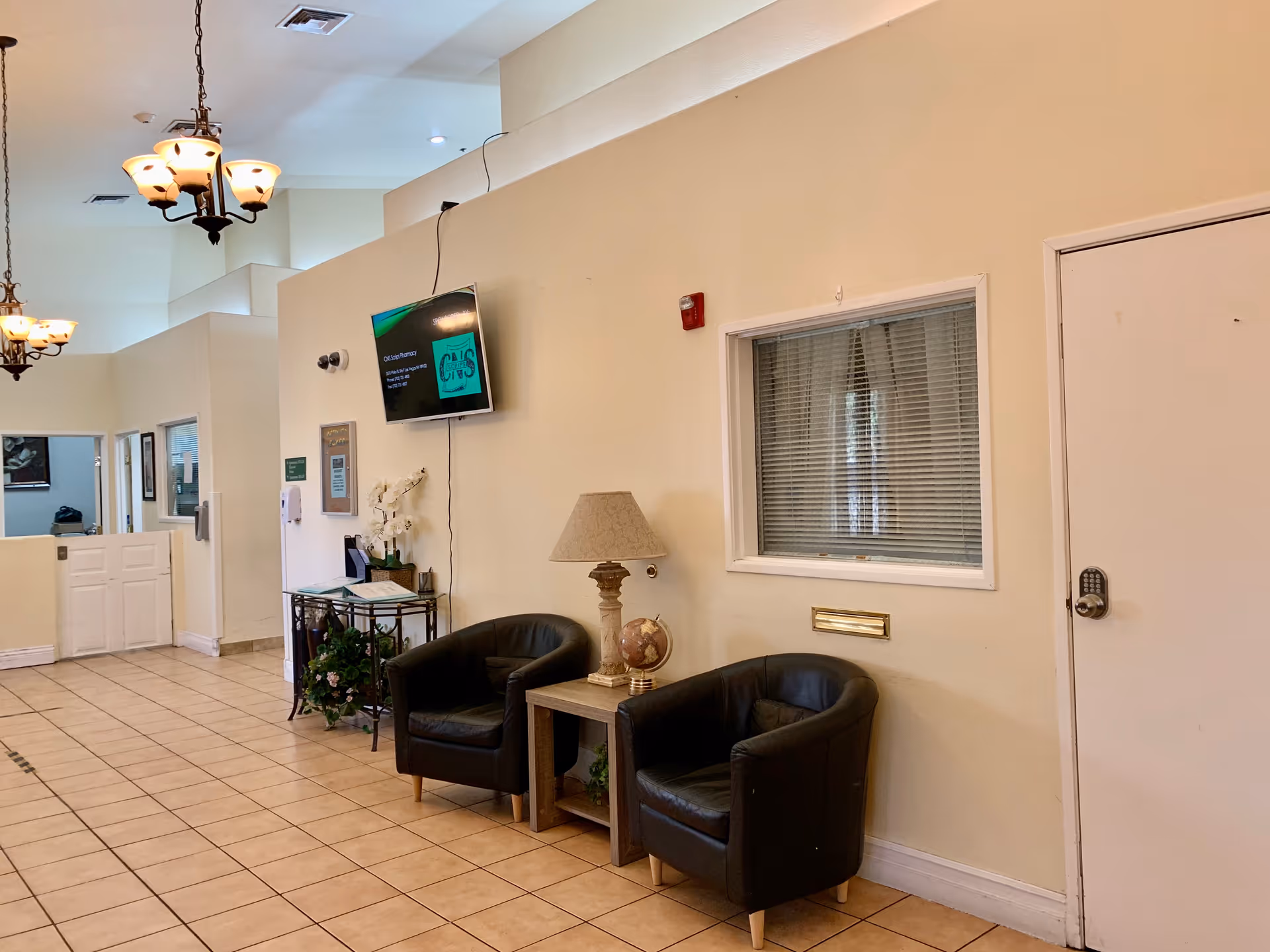 A waiting area inside Sana Assisted Living with two black armchairs and a small wooden table between them holding a lamp and a globe. A wall-mounted TV is above a small table with flowers and informational materials. The floor is tiled, and there are hanging light fixtures on the ceiling. A window with blinds and a door with a keypad lock are visible on the right wall.