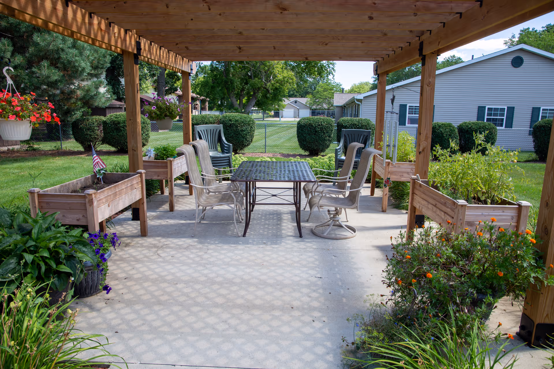 Outdoor covered patio area with a wooden pergola overhead, featuring a metal table surrounded by six chairs. Raised garden beds with various plants and flowers are placed around the patio. In the background, there is a green lawn, trimmed bushes, trees, and a building with gray siding and green shutters.