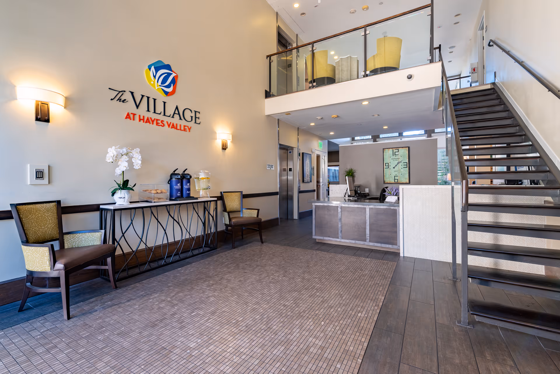 Lobby area of The Village at Hayes Valley featuring a reception desk, staircase leading to an upper floor with seating, two chairs and a table with refreshments including water and coffee, and a wall sign with the facility's name and logo.