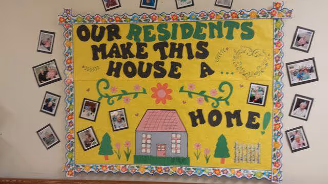 A colorful bulletin board with the message 'Our residents make this house a home' surrounded by small photos of residents. The board is decorated with flowers, a house drawing, trees, and a fence.