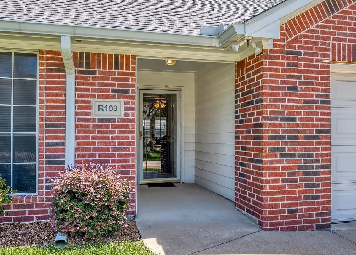 Entrance to a residential unit labeled R103 at Reunion Inn Assisted Living, featuring a brick exterior, a glass front door with a white frame, a small porch area, and a bush with pink and green leaves near the entrance.