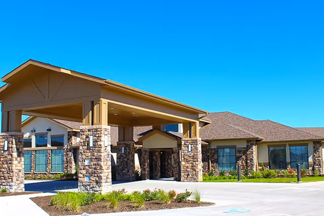 Exterior view of a single-story senior living facility building with a covered entrance supported by stone pillars, surrounded by landscaped greenery under a clear blue sky.
