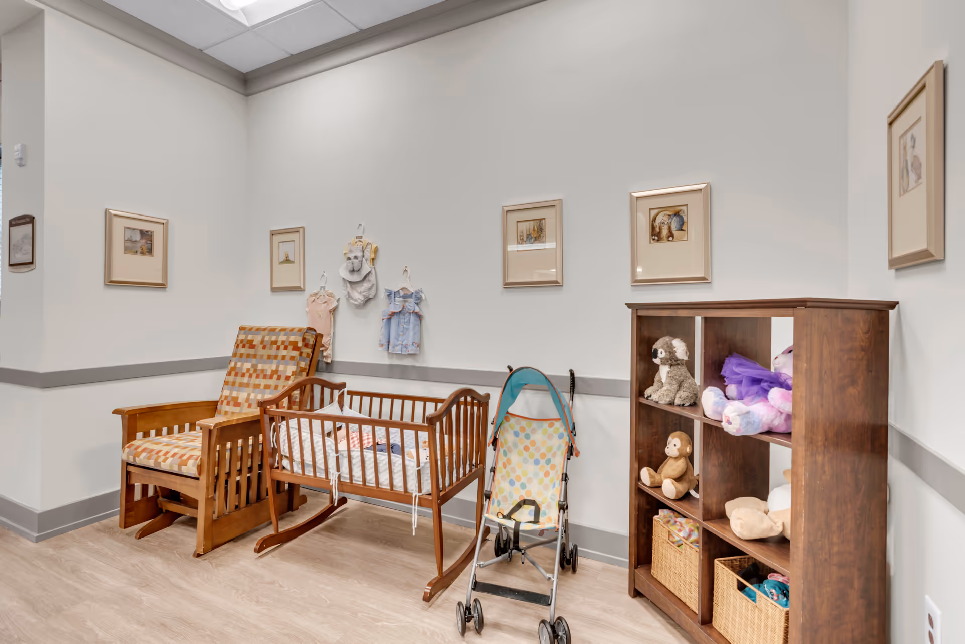 A cozy corner of a room featuring a wooden rocking chair with patterned cushions, a wooden cradle with bedding, a small stroller, and a wooden cubby shelf filled with stuffed animals and baskets. The walls are decorated with framed pictures and baby clothes hanging on hooks.