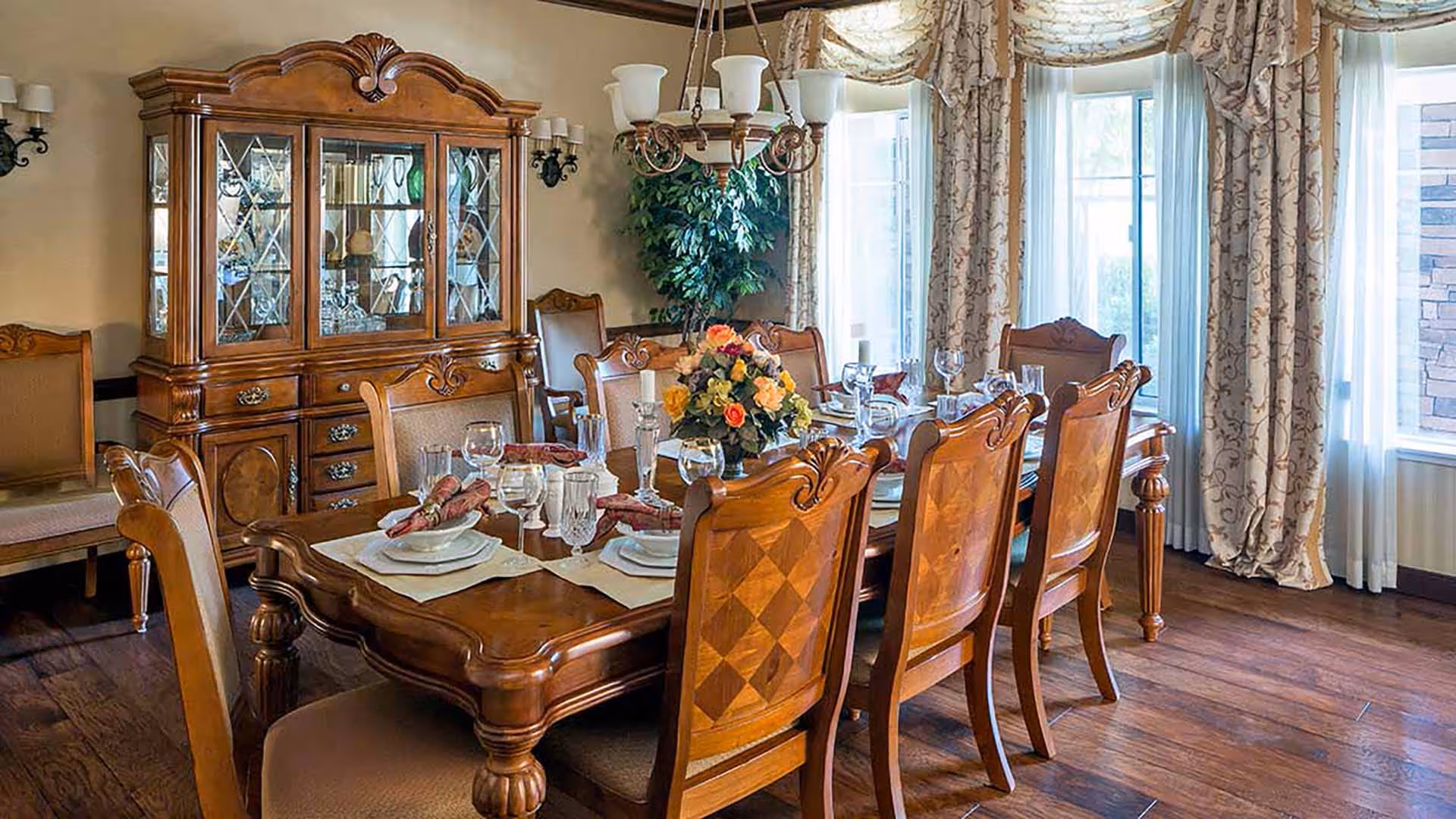 A formal dining room with an ornate wooden dining table set for six, featuring plates, glasses, and napkins. The room has matching wooden chairs with cushioned seats, a large wooden china cabinet with glass doors displaying dishes, a chandelier hanging above the table, floral curtains on the windows, and a potted plant in the corner.