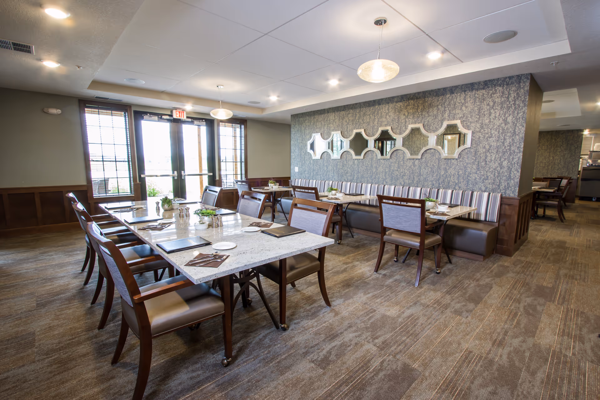 A bright dining room with multiple tables and chairs arranged neatly. The tables have place settings including plates, napkins, and menus. There is a long cushioned bench along one wall with striped upholstery, and decorative mirrors hang above it. Large windows and a glass door allow natural light to fill the room. The floor is carpeted and the ceiling has recessed lighting and pendant lights.